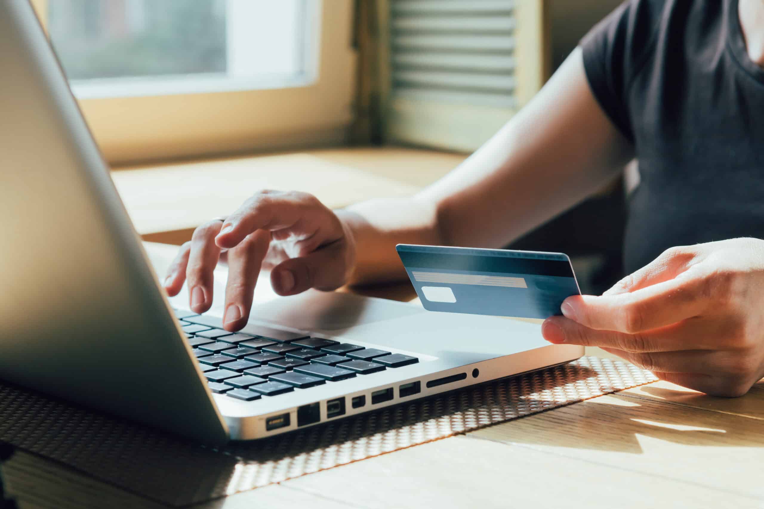 A person holding a credit card in one hand and using the other hand to type on a laptop keyboard, sitting at a table near a window with sunlight coming in.
