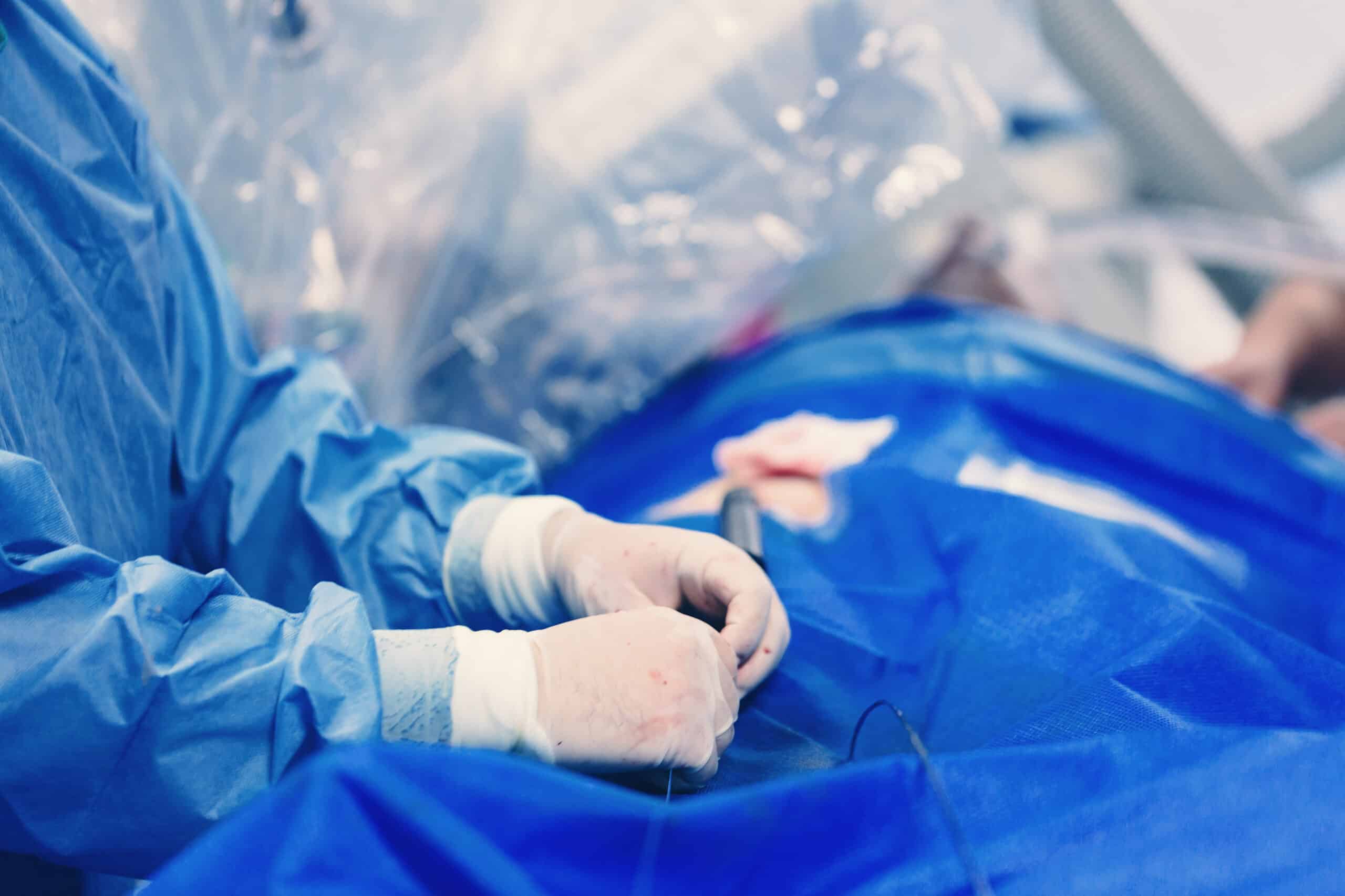 A medical professional wearing blue surgical scrubs and gloves performs a procedure on a patient covered with a blue sterile drape in an operating room.