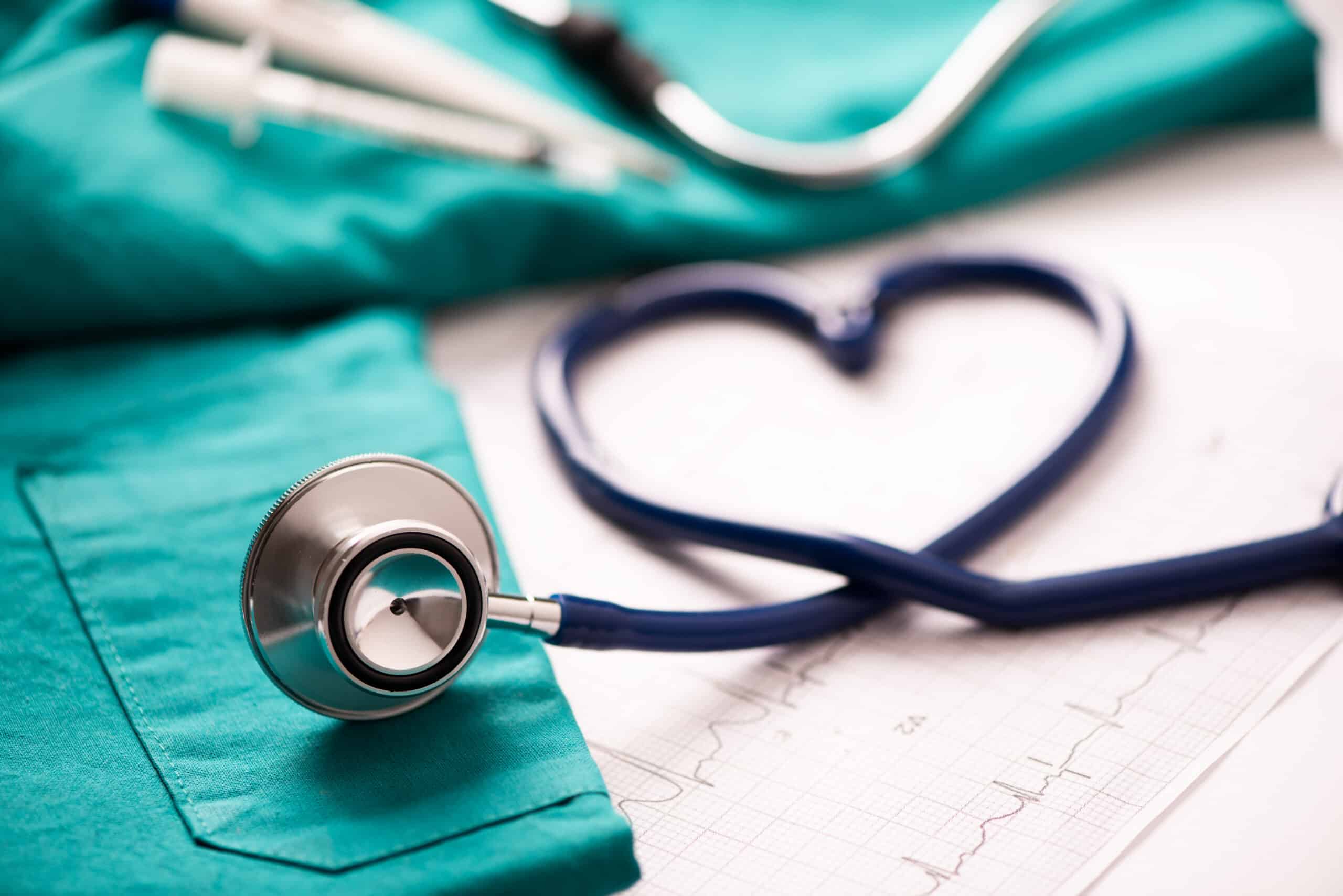 A stethoscope resting on green scrubs with its tubing shaped like a heart on top of an electrocardiogram ECG printout, symbolizing healthcare and compassion.
