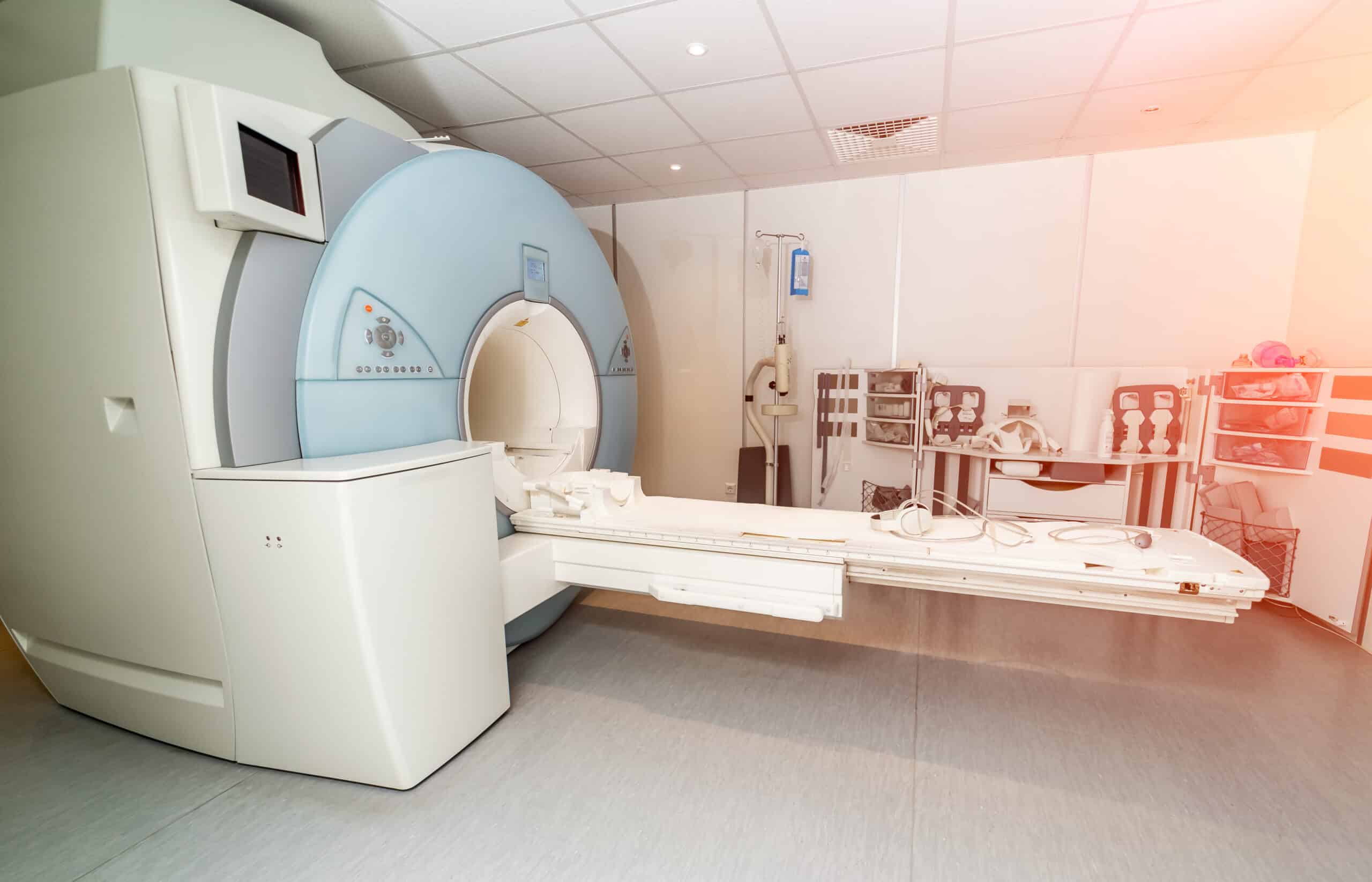 A large MRI scanner in a clean, brightly lit medical room with an extended patient table and various medical equipment and control panels nearby.