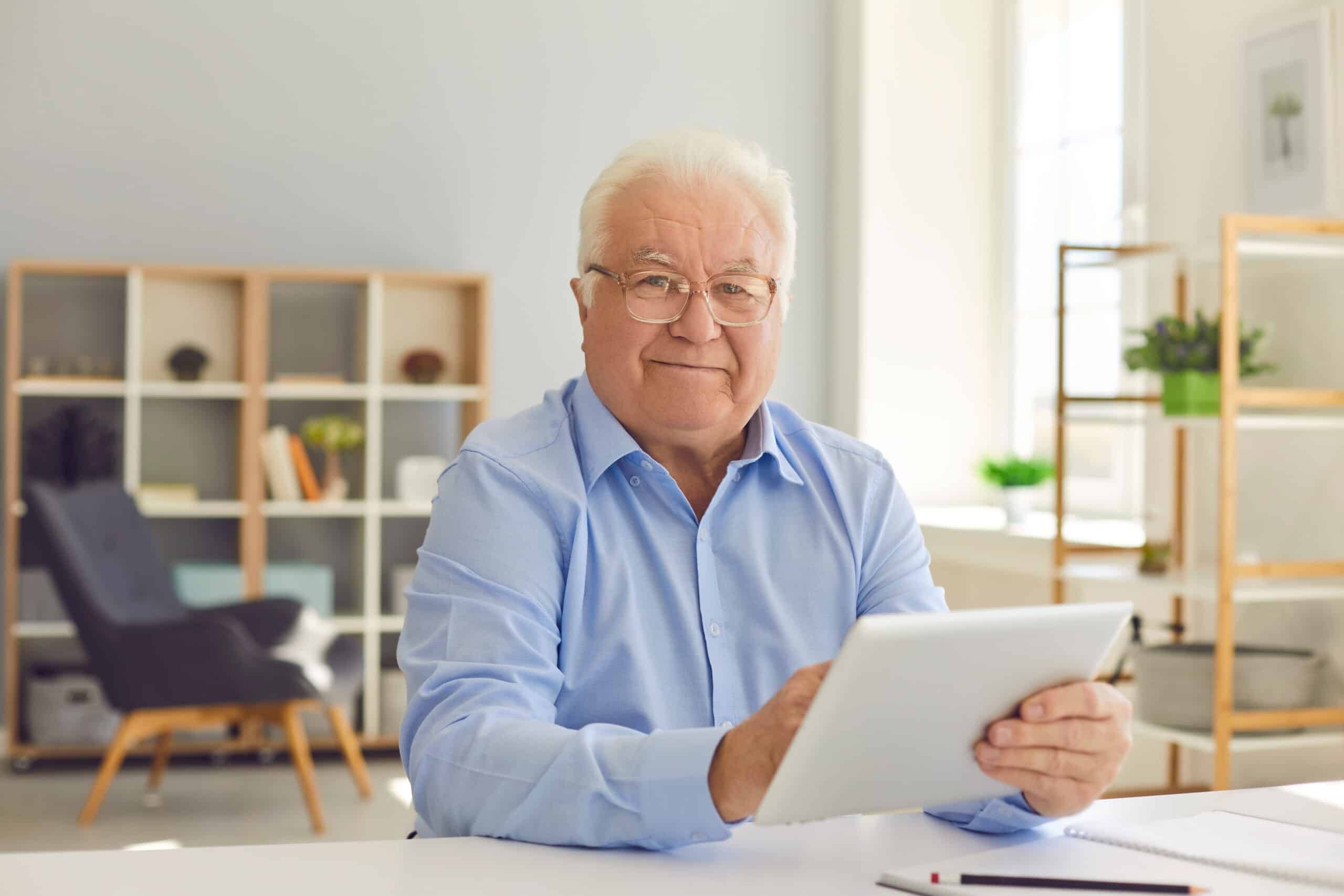 An elderly man with white hair and glasses smiles while holding a tablet at a desk in a bright, modern home office with shelves and plants in the background.