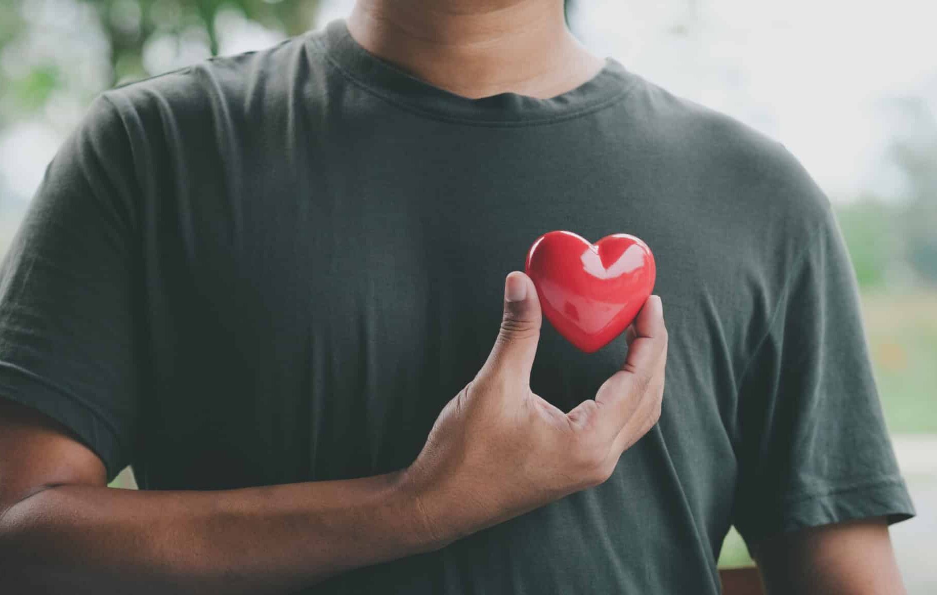 A person wearing a dark green t-shirt holds a red heart-shaped object against their chest, symbolizing care or love. The background is blurred with greenery.