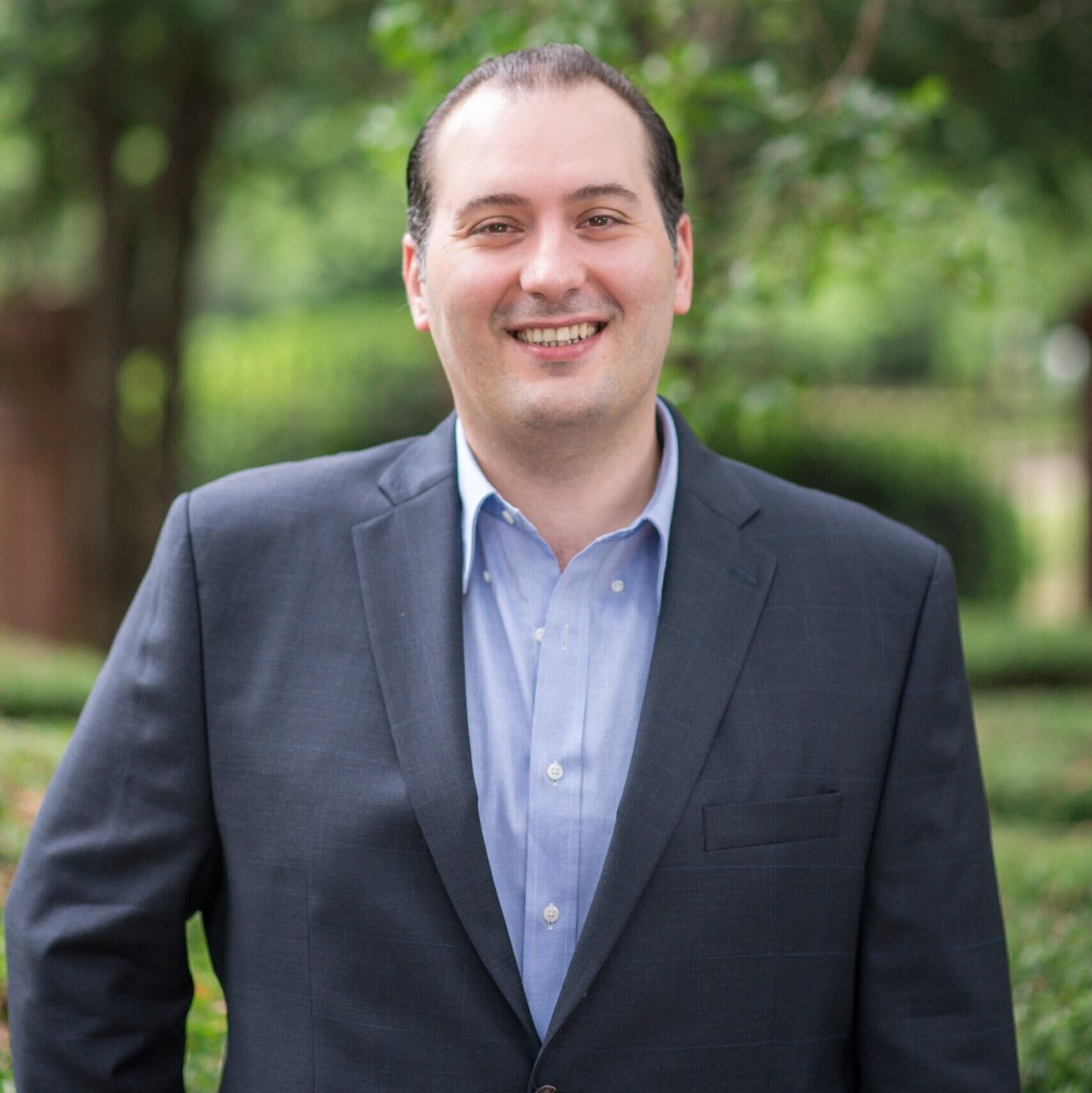 A man in a dark suit and light blue shirt stands outdoors, smiling, with green trees and foliage blurred in the background.