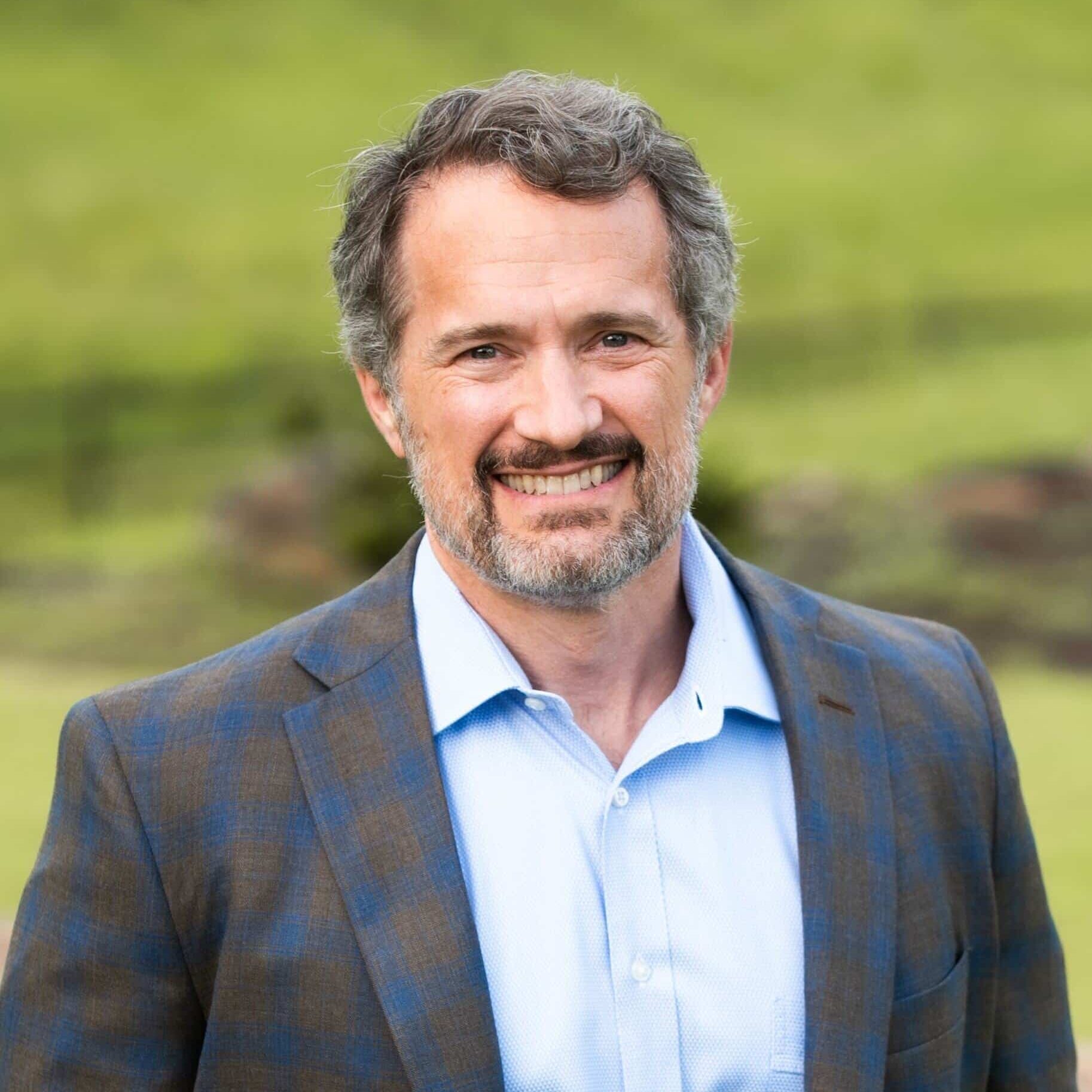 A smiling middle-aged man with gray hair and a beard, wearing a blue dress shirt and a checked blazer, stands outdoors in front of a blurred green background.