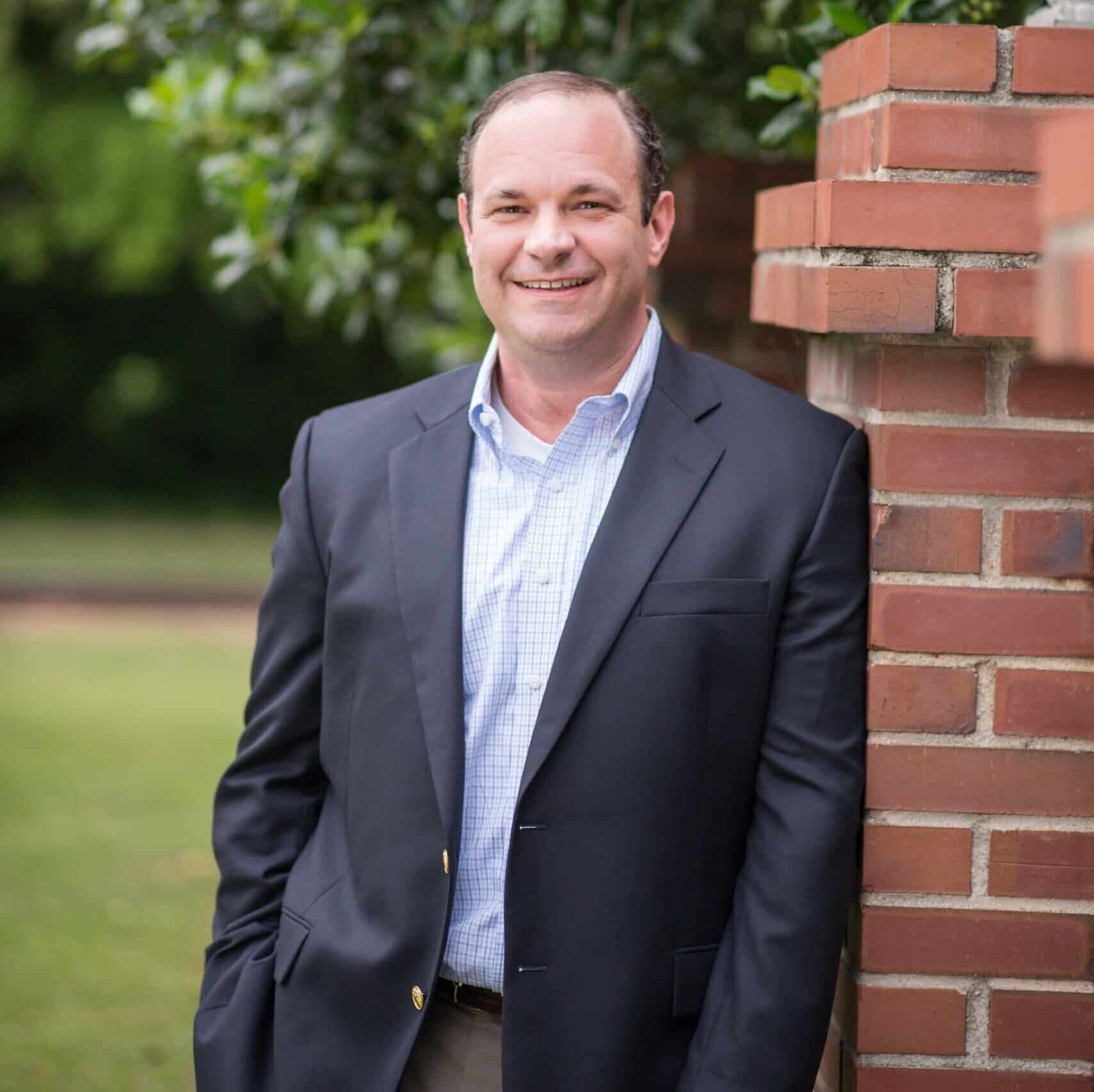 A man in a navy blazer and checked shirt smiles while leaning against a brick wall outside, with green trees and leaves in the background.
