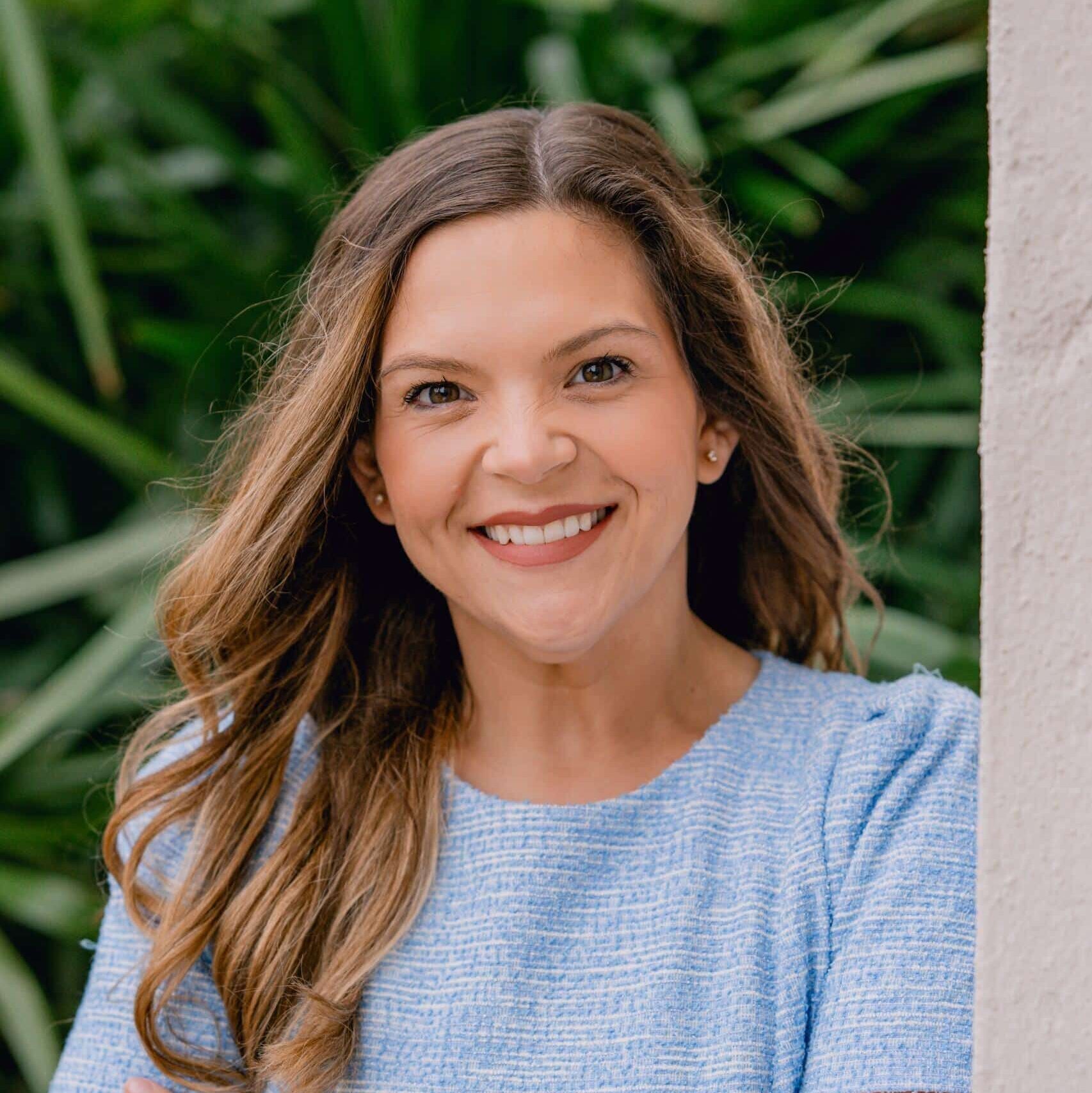 A woman with long wavy brown hair, wearing a light blue textured short-sleeve top, smiles with arms crossed while standing in front of green foliage and a white wall.