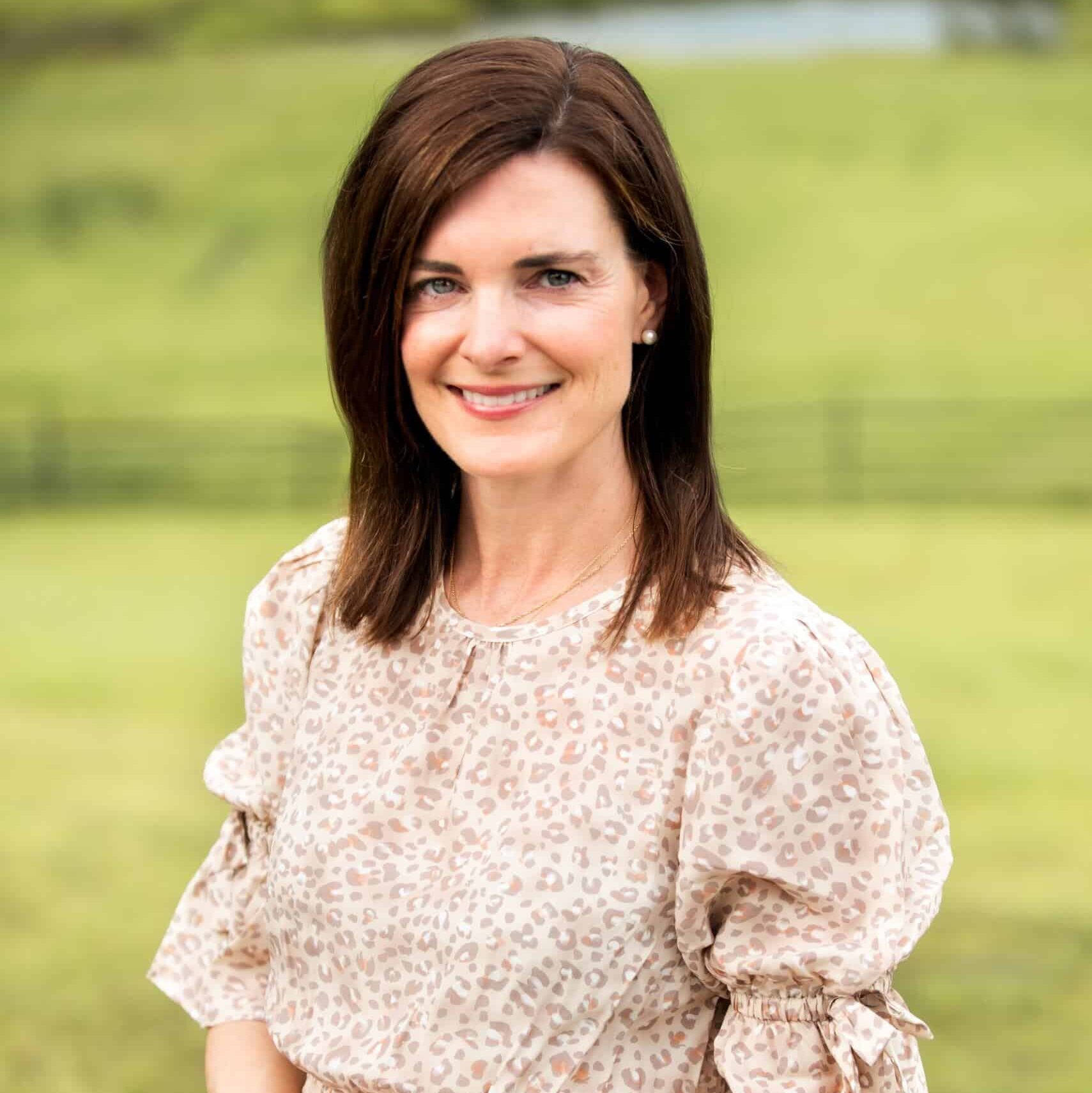 A woman with shoulder-length brown hair, wearing a light-colored long-sleeve dress with a subtle leopard print, stands outdoors on a grassy field, smiling at the camera.