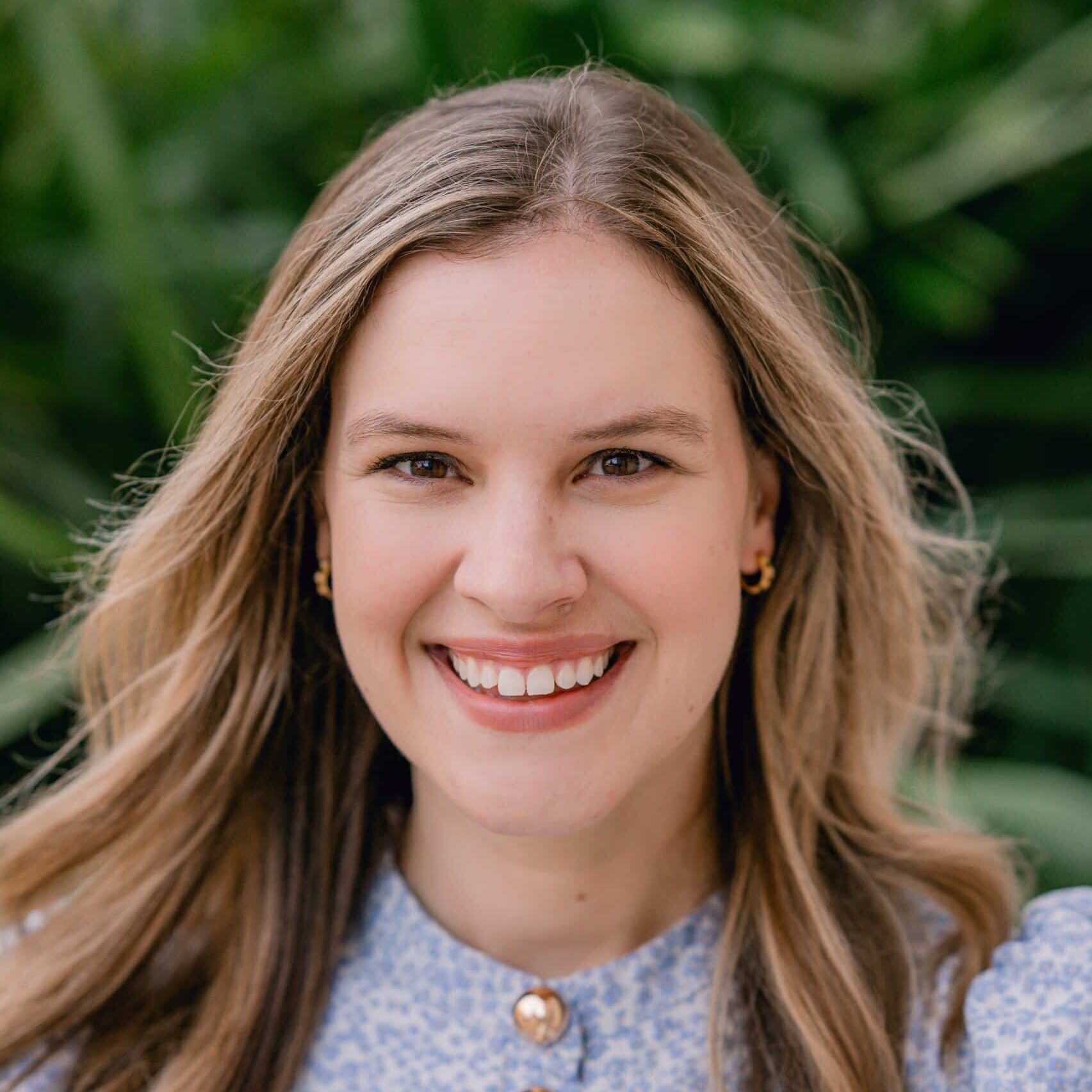 A woman with long light brown hair smiles at the camera. She is wearing a light blue patterned blouse with gold buttons. The background is blurred greenery.