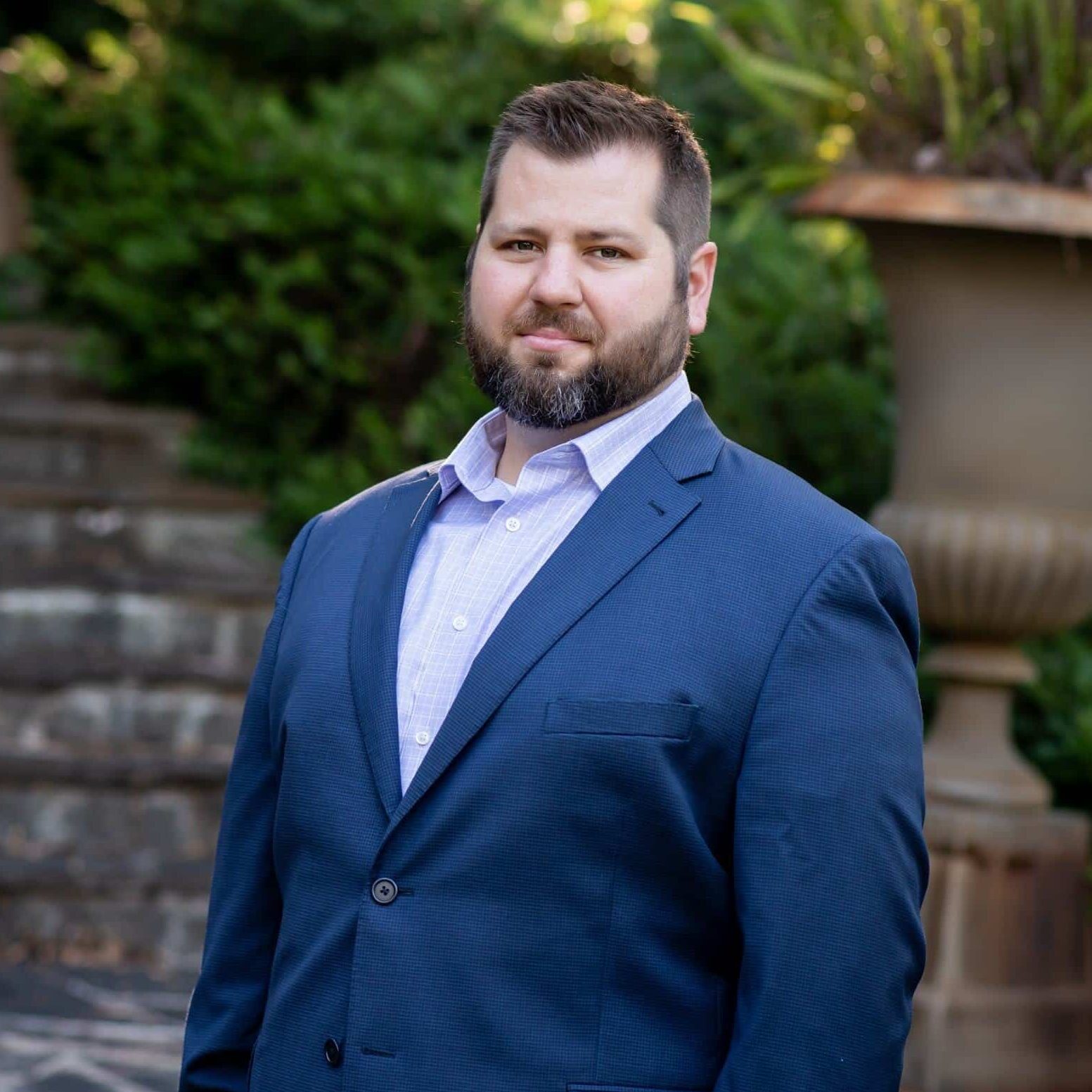 A man with a short beard and dark hair, wearing a blue blazer, light blue shirt, and tan pants, stands outdoors in front of stone steps and greenery, looking confidently at the camera.