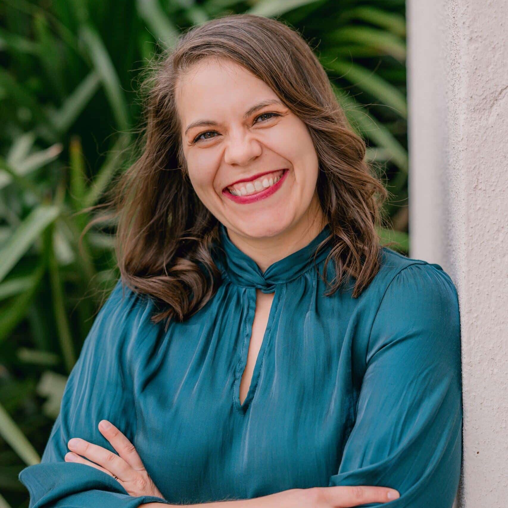 A woman with shoulder-length brown hair and a bright smile stands with arms crossed, wearing a teal blouse, in front of green foliage and a white wall.