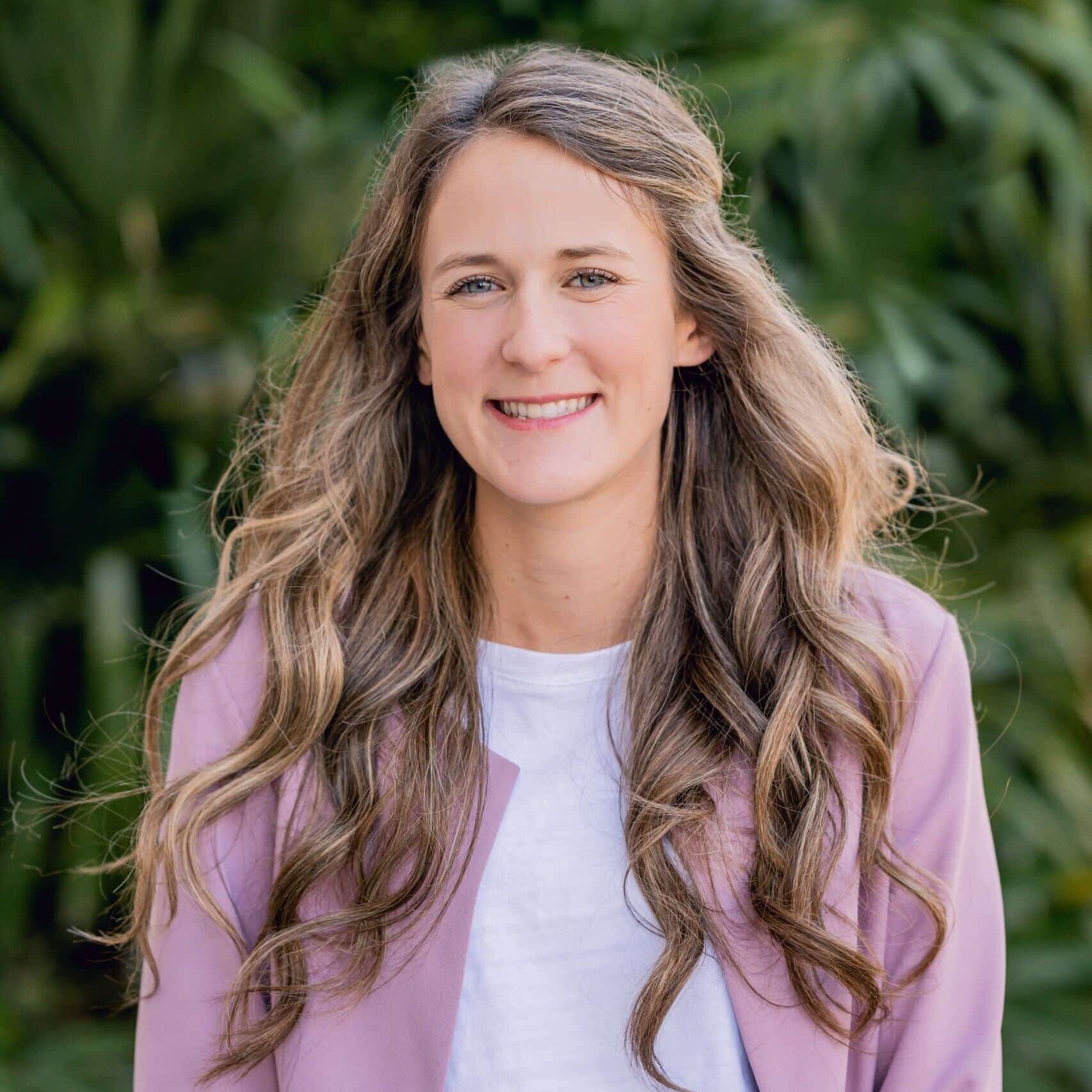 A woman with long, wavy light brown hair smiles at the camera. She is wearing a white shirt and a light purple blazer. The background is filled with green, leafy plants.