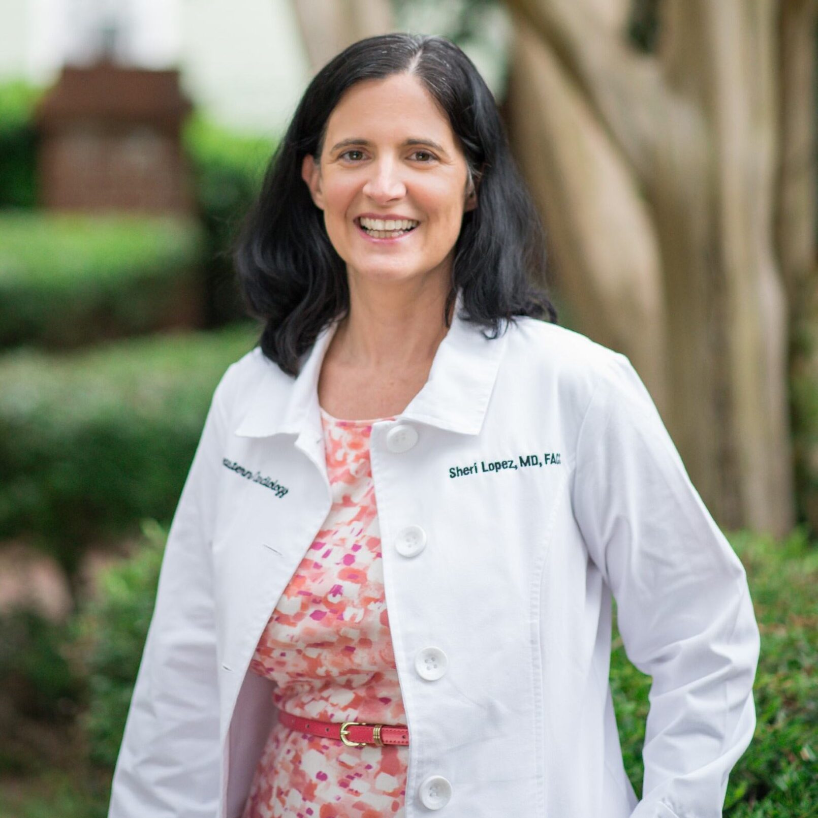 A woman with dark hair wearing a white lab coat and a pink patterned dress stands outside, smiling, with trees and greenery in the background.