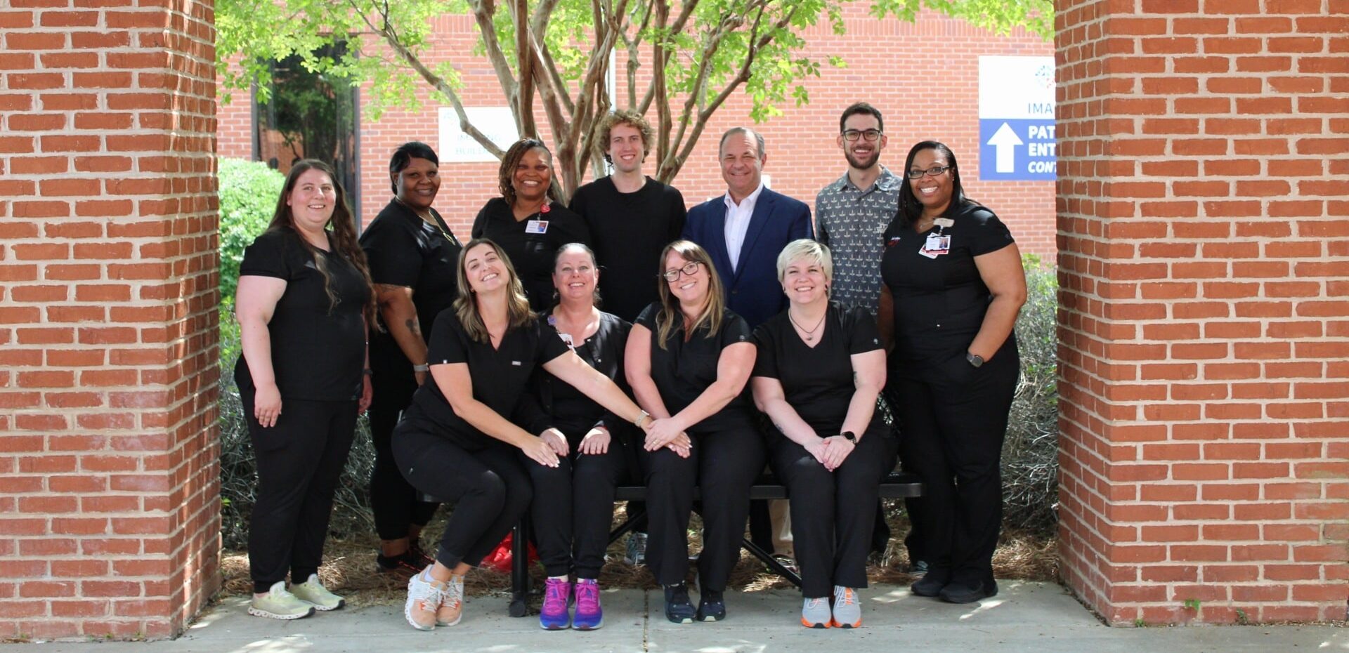 A group of twelve people, mostly in black uniforms, pose and smile outdoors under a tree between two brick columns. Some are seated on a bench while others stand behind them. A brick building is visible in the background.