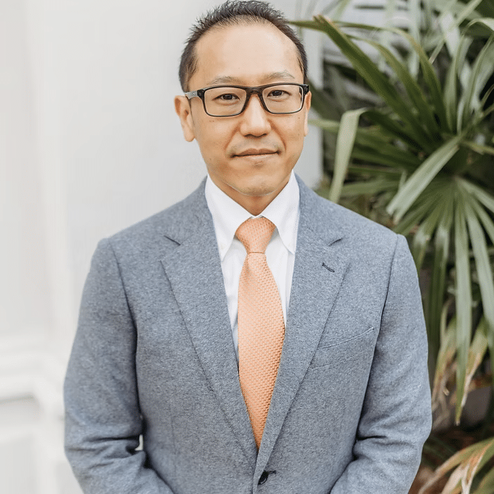 A man wearing glasses, a grey suit, white shirt, and peach-colored tie stands in front of a white wall and green leafy plant, smiling slightly at the camera.