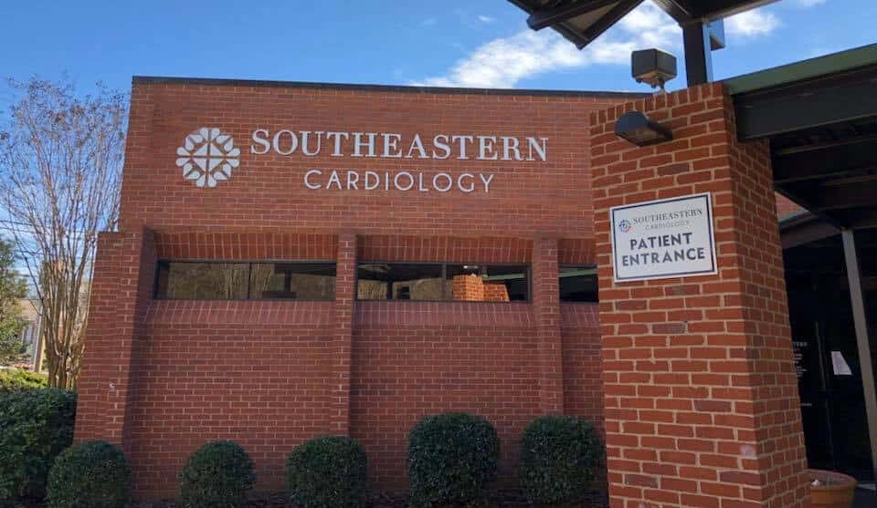 A brick building with a sign reading Southeastern Cardiology. A smaller sign near the entrance says Patient Entrance. Trees and bushes surround the building on a clear day.