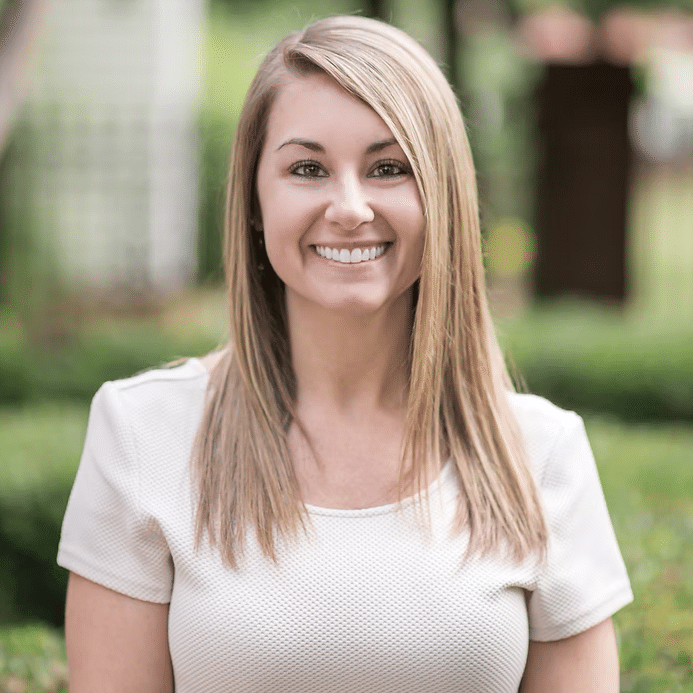 A young woman with straight, light brown hair and a beige short-sleeved top smiles while standing outdoors in a garden with blurred greenery in the background.