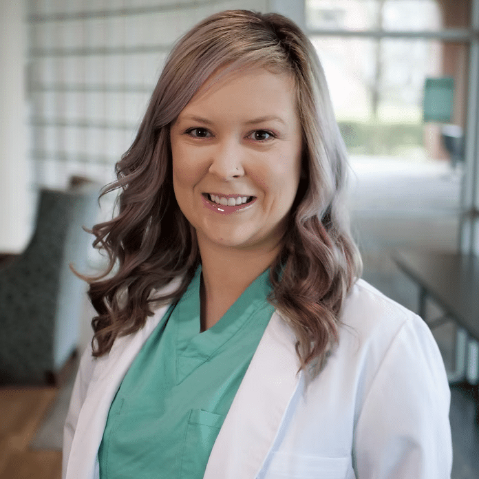 A woman wearing a white lab coat over green scrubs smiles at the camera in a bright, modern indoor setting with large windows and furniture in the background.