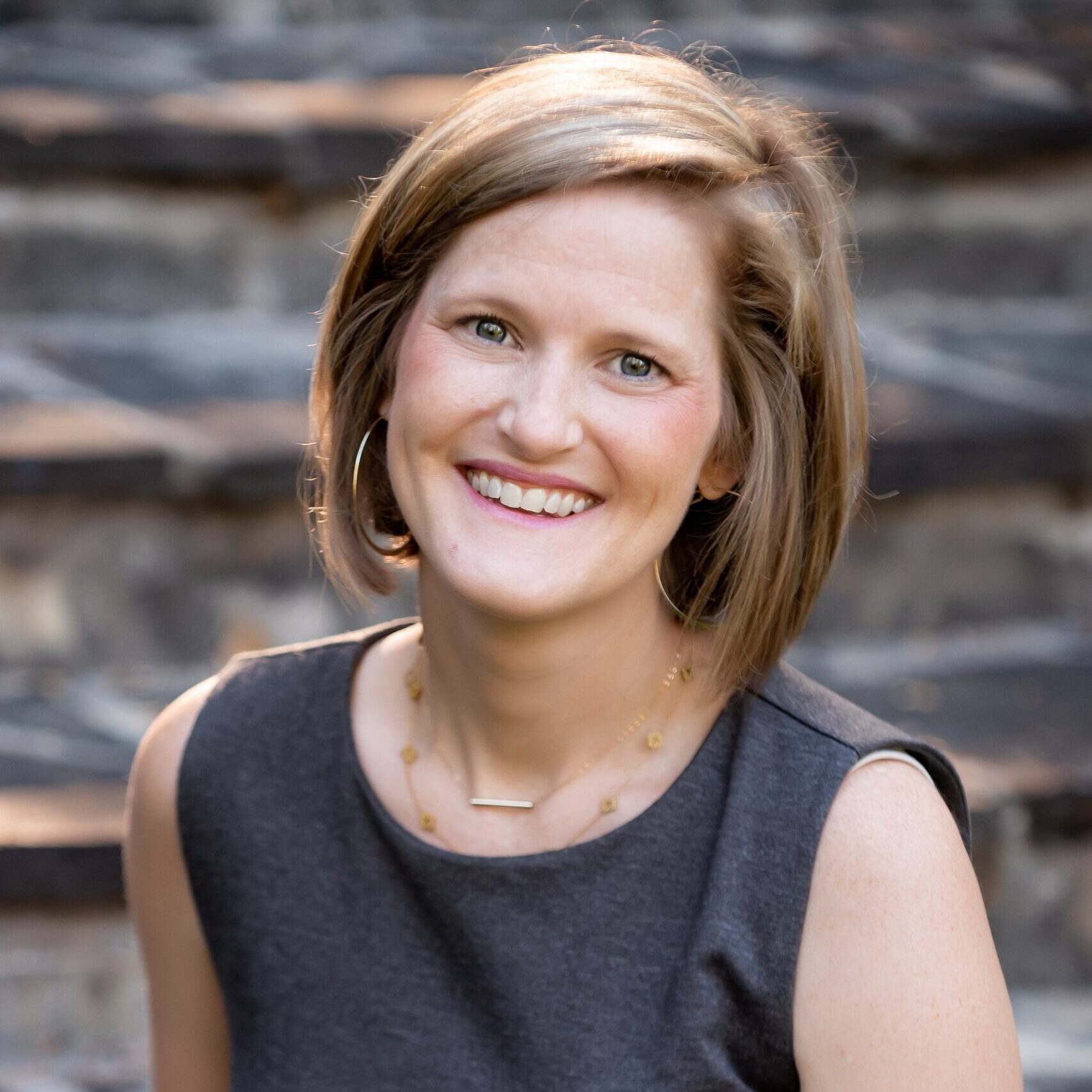 A woman with short light brown hair, wearing a sleeveless dark gray dress and a delicate necklace, smiles while sitting outdoors in front of stone steps.