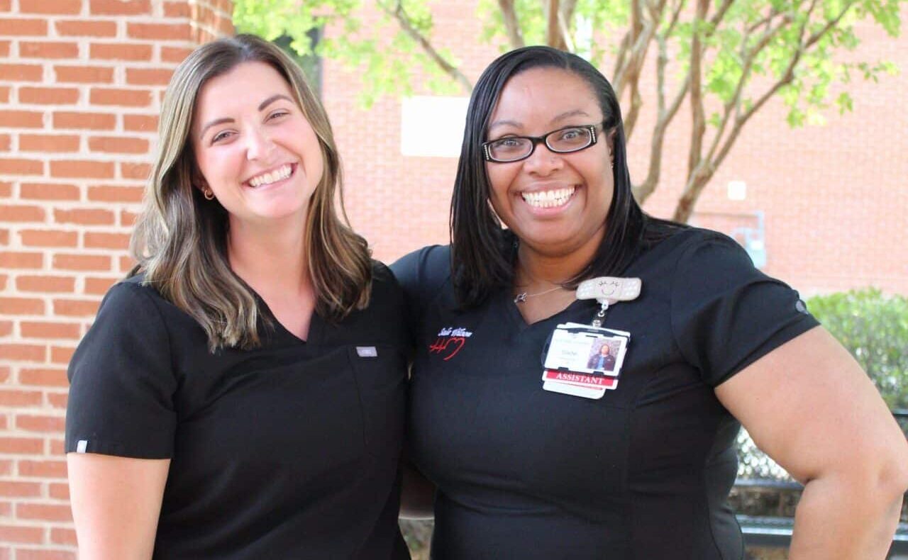 Two women in black medical scrubs smile while standing outside in front of a brick wall and green trees. One woman has a name badge and glasses, and both appear happy and relaxed.