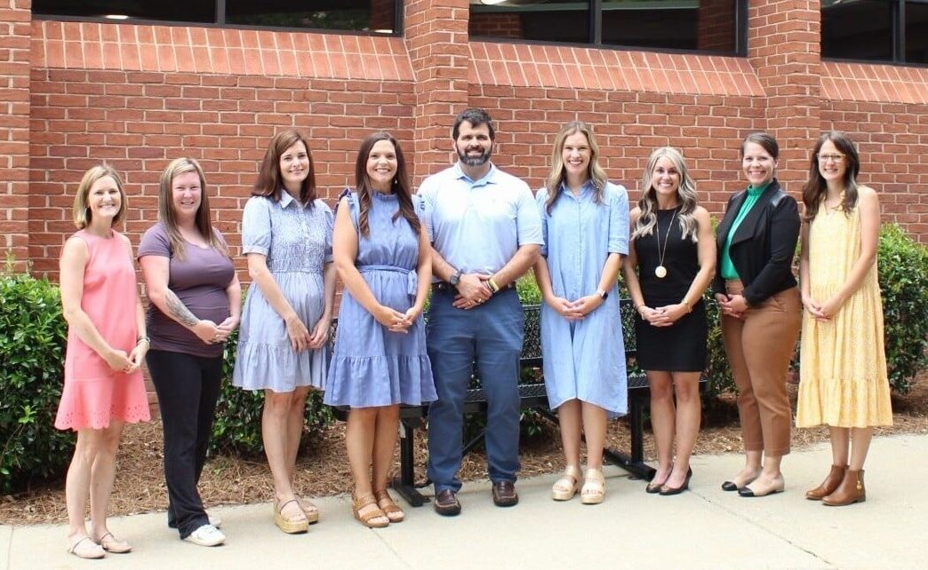 Ten people stand in a row smiling outside a brick building with a sign that reads Southeastern Cardiology. Shrubs line the walkway in front of the building.