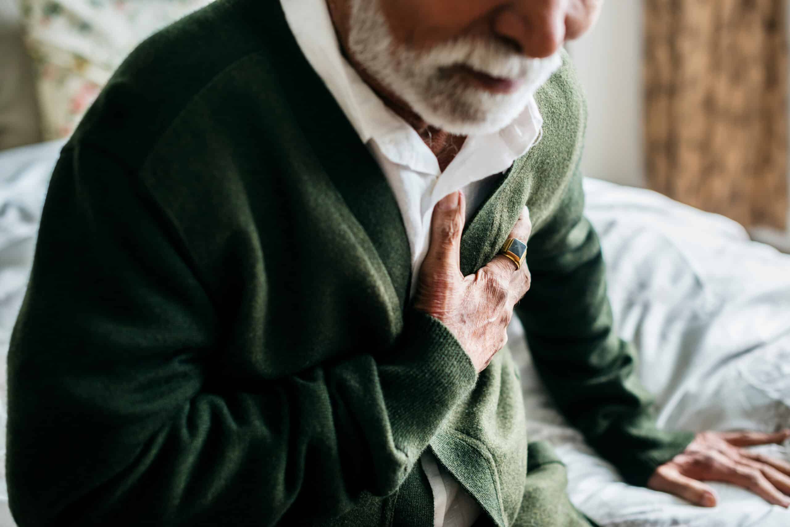 An older man with a white beard in a green cardigan and white shirt sits on a bed, clutching his chest with a look of pain.