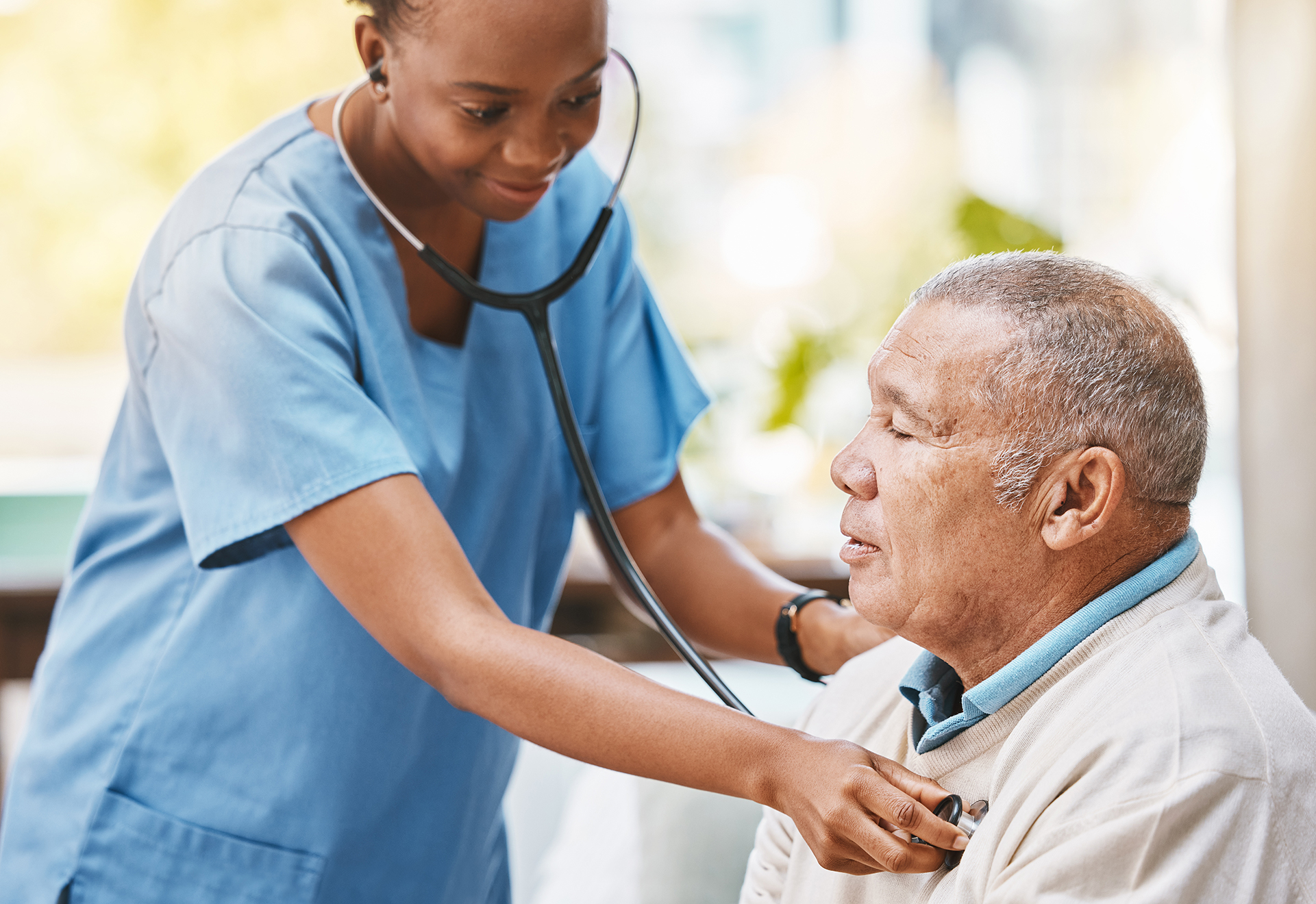 A nurse in blue scrubs listens to the chest of an elderly man in a beige sweater with a stethoscope during a check-up.