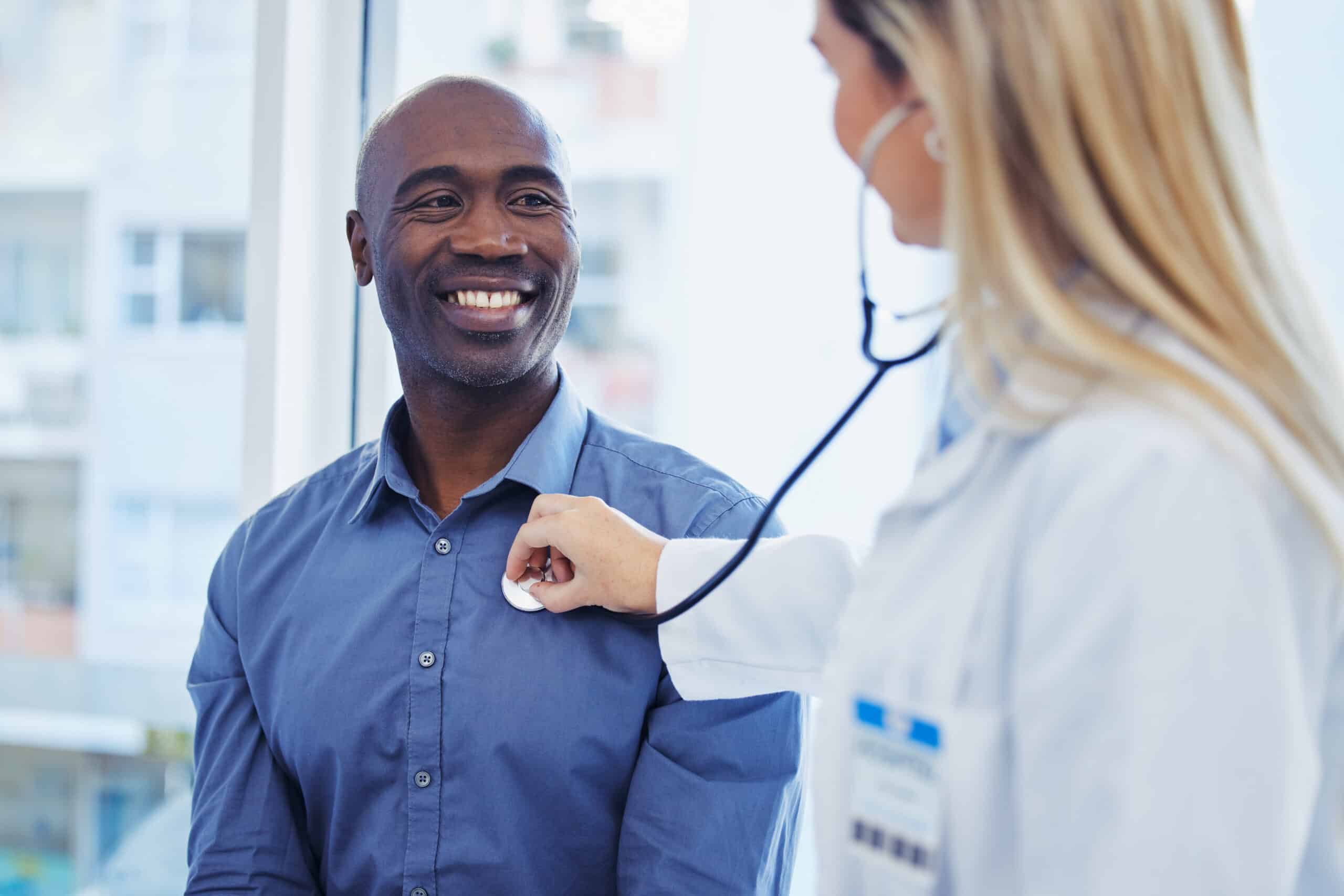 A smiling man sits as a healthcare professional listens to his chest with a stethoscope during a checkup in a bright office.