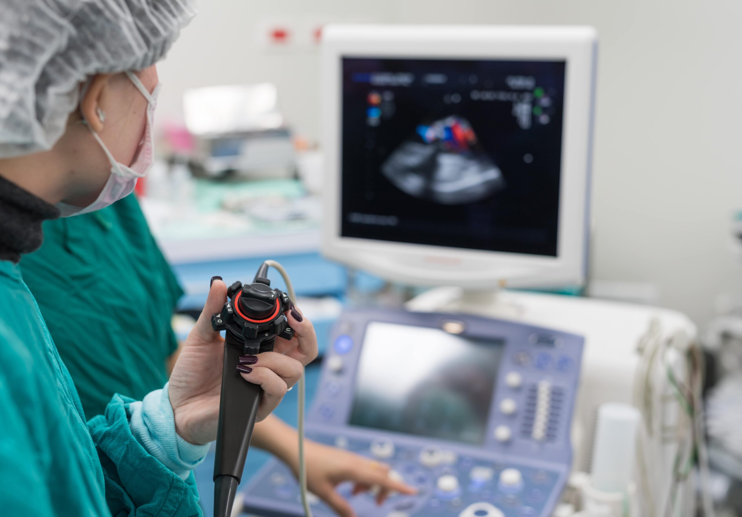 A masked medical professional with a hair cover holds an endoscope as another operates an ultrasound showing colored images in a clinic.