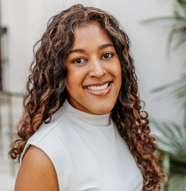 A woman with long, curly hair and a warm smile wears a light sleeveless top, standing outside near green plants, facing the camera.