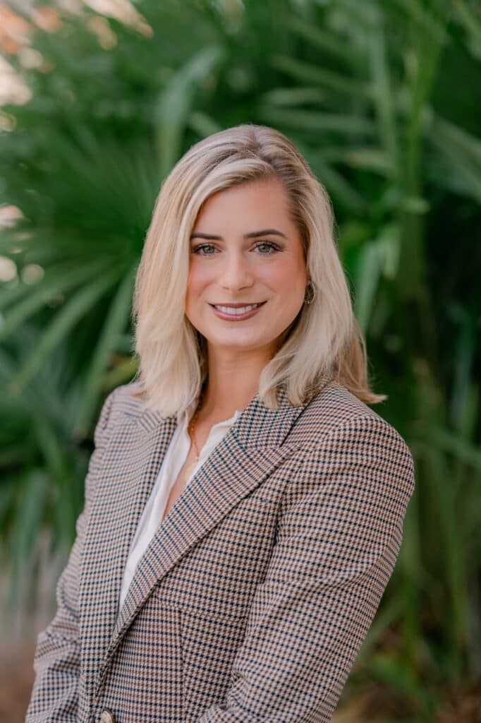 Blonde-haired woman in a brown checkered blazer and white shirt smiles outdoors, standing before lush green foliage.