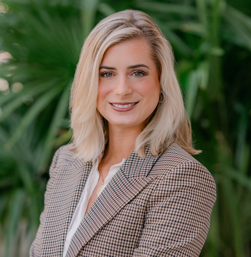Blonde-haired woman in a brown checkered blazer and white shirt smiles outdoors, standing before lush green foliage.